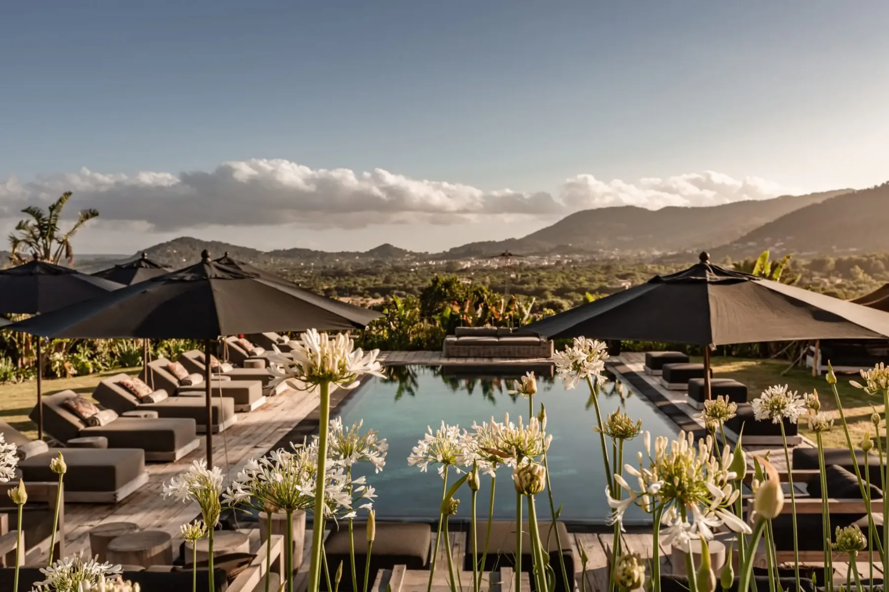 Luxurious outdoor pool with black umbrellas, sun loungers, and white flowers, set against a backdrop of rolling hills and a clear sky.