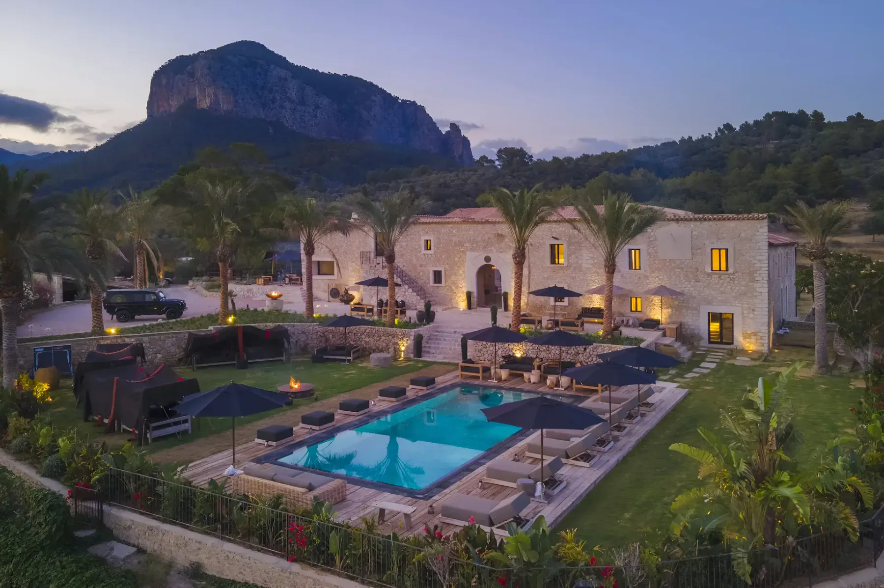 Luxurious villa at dusk with a lit swimming pool, surrounded by palm trees and mountains in the background.