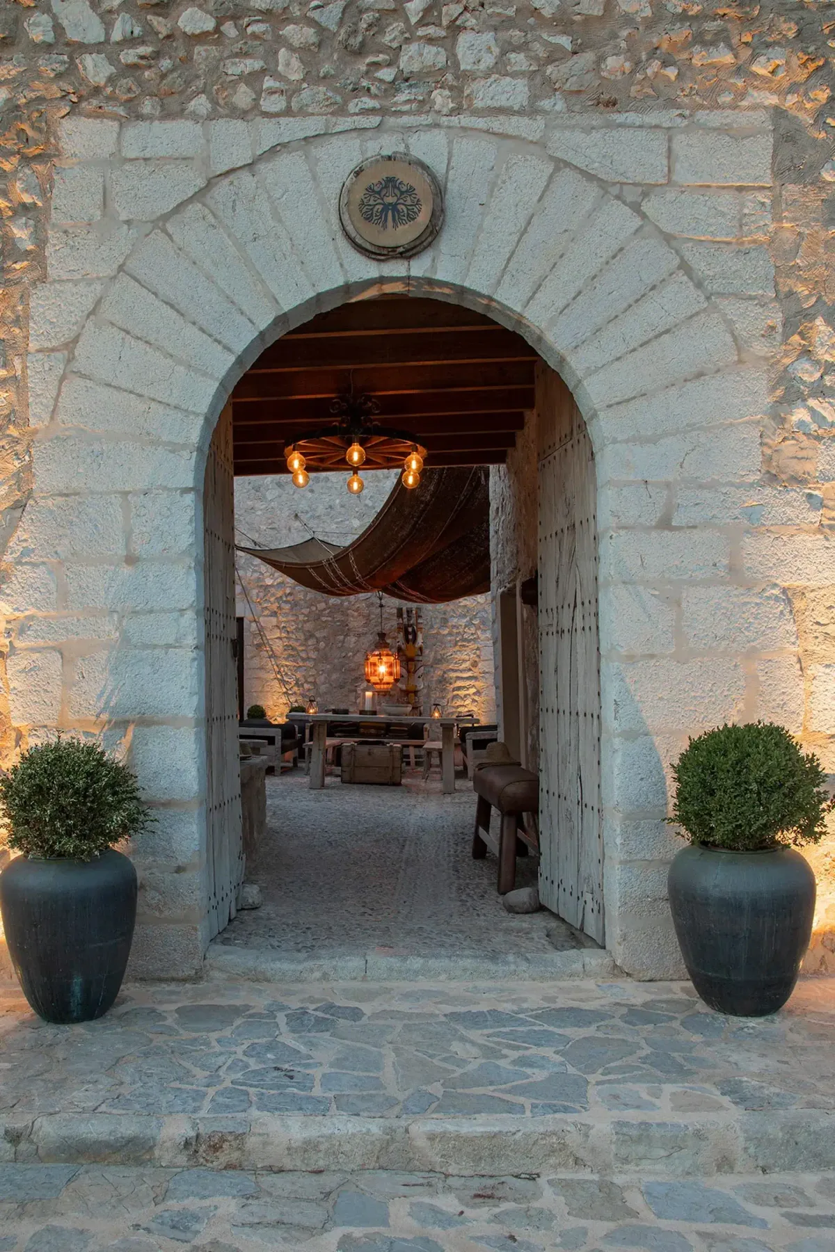 Stone archway entrance with rustic decor, two potted plants, warm lighting, and a hanging chandelier leading to an outdoor seating area.