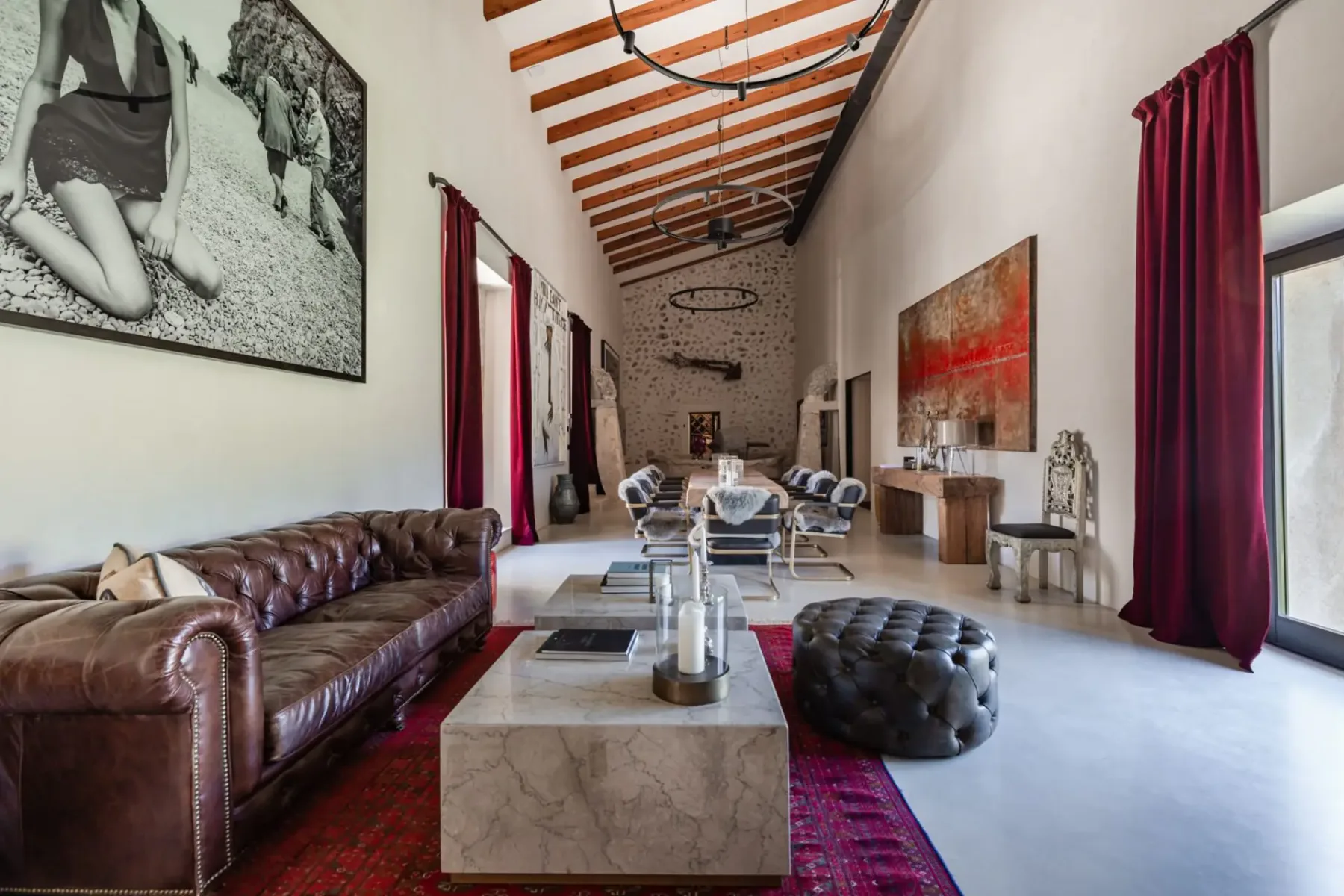 Spacious living room with leather sofa, modern art, marble coffee table, and wooden beams. Red curtains and large windows add elegance.