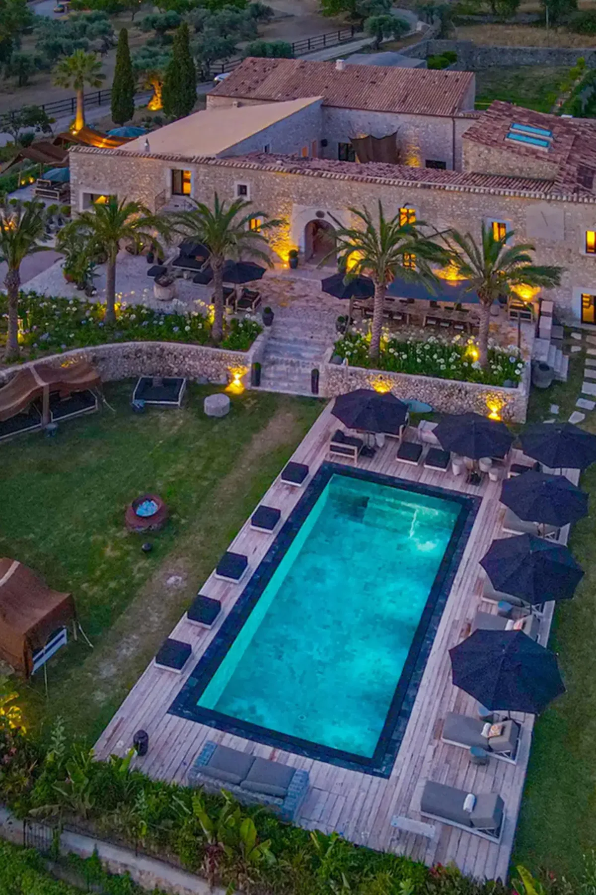 Aerial view of a luxurious villa with stone walls, a lit swimming pool, palm trees, and patio seating, surrounded by lush greenery.