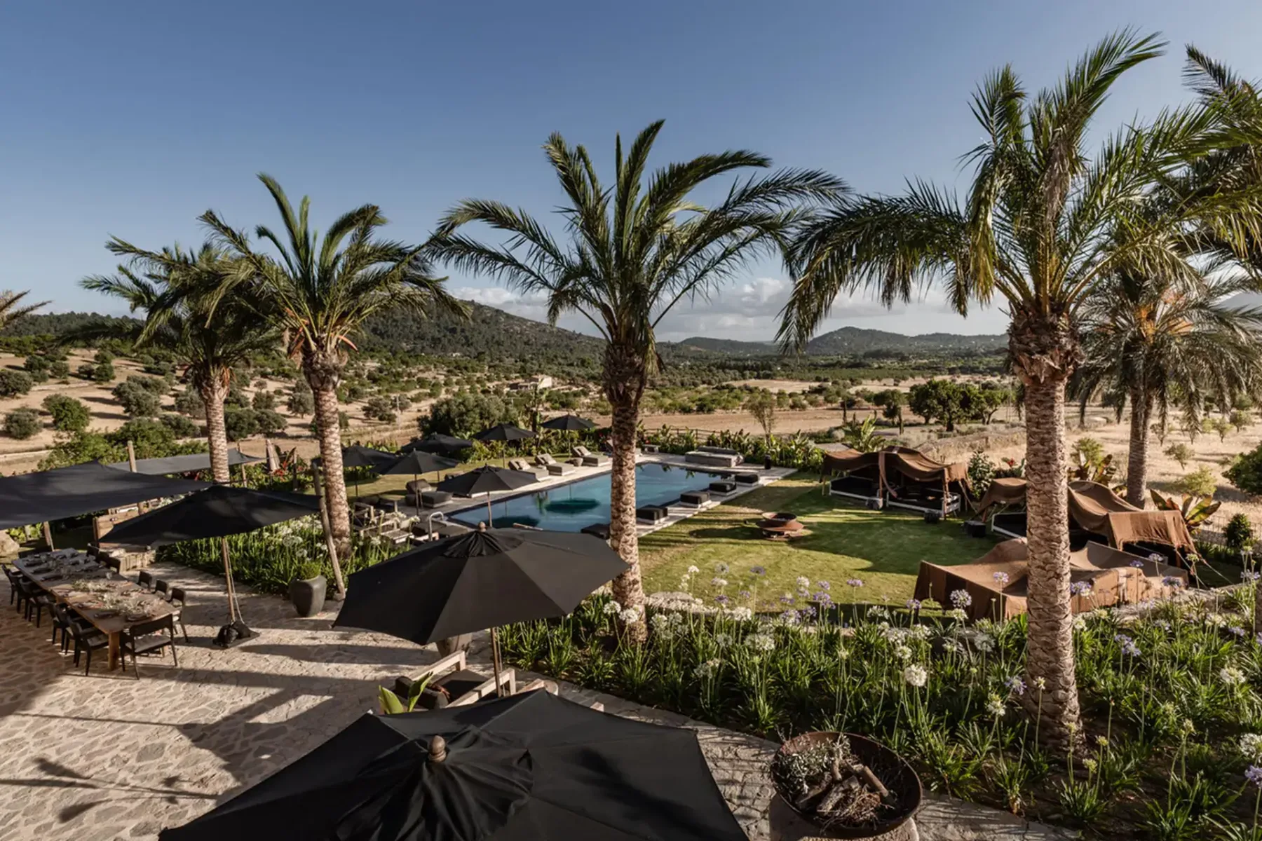 A luxurious outdoor area with a pool, surrounded by palm trees and umbrellas, set in a desert landscape with distant mountains.