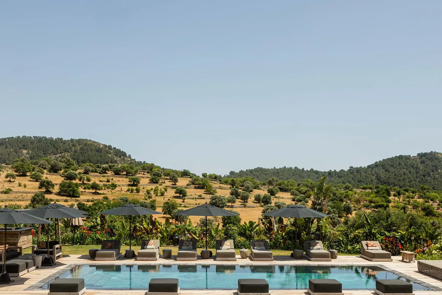 Outdoor pool with sun loungers and umbrellas, surrounded by lush greenery and rolling hills under a clear blue sky.