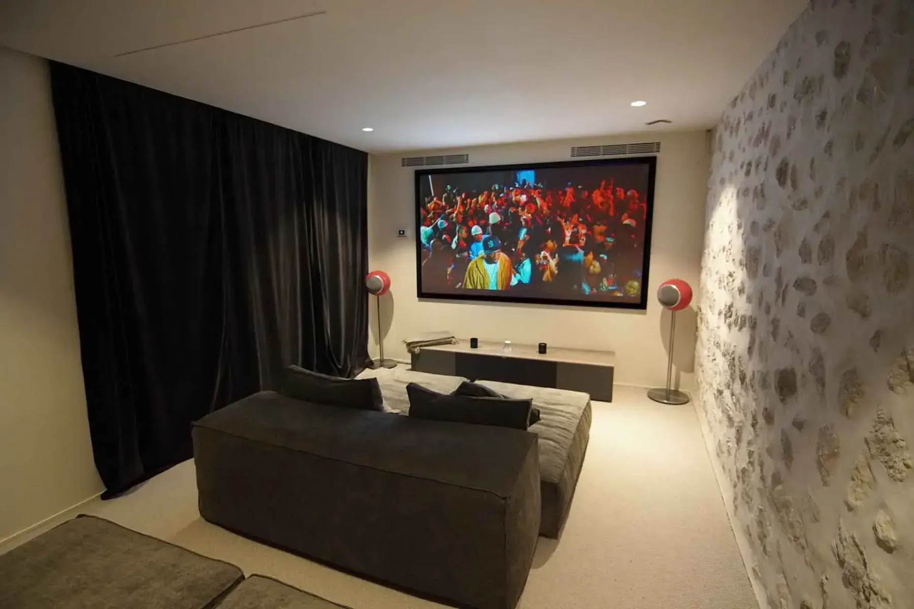 Home theater room with a large screen showing a concert, two gray sofas, light stone wall, and dark curtains.