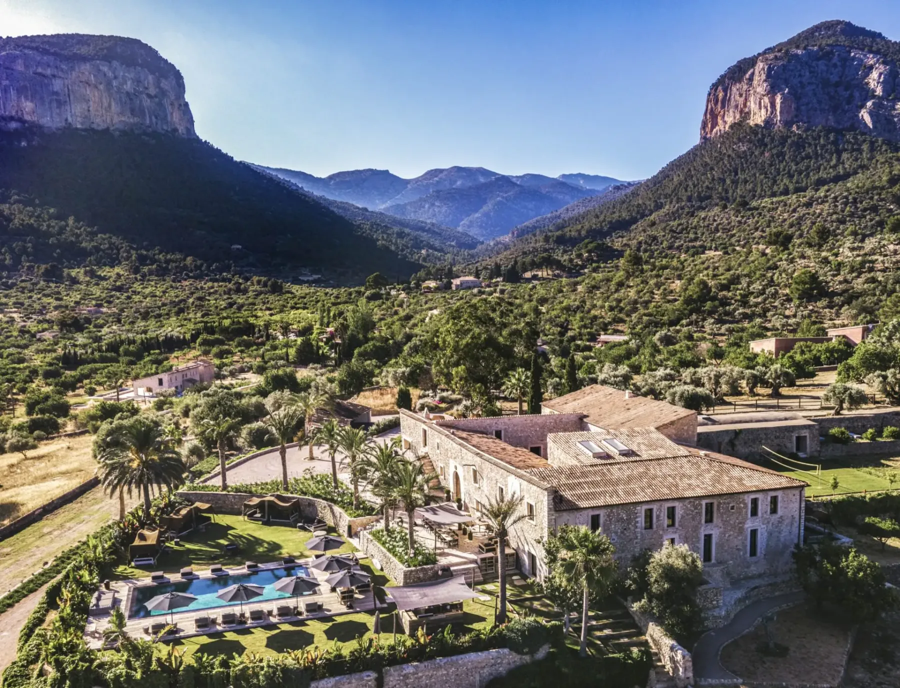 Aerial view of a large stone villa with a swimming pool, surrounded by lush greenery and mountains under a clear blue sky.