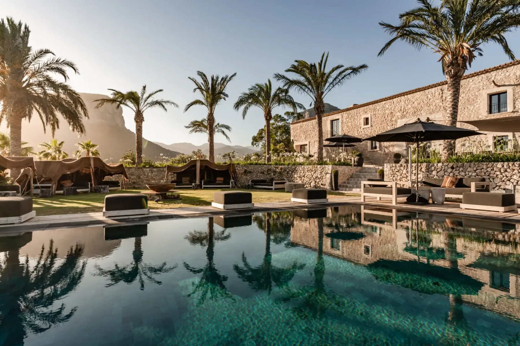 Luxurious stone villa with a reflective pool, surrounded by palm trees and sun loungers, set against a mountainous landscape at sunset.