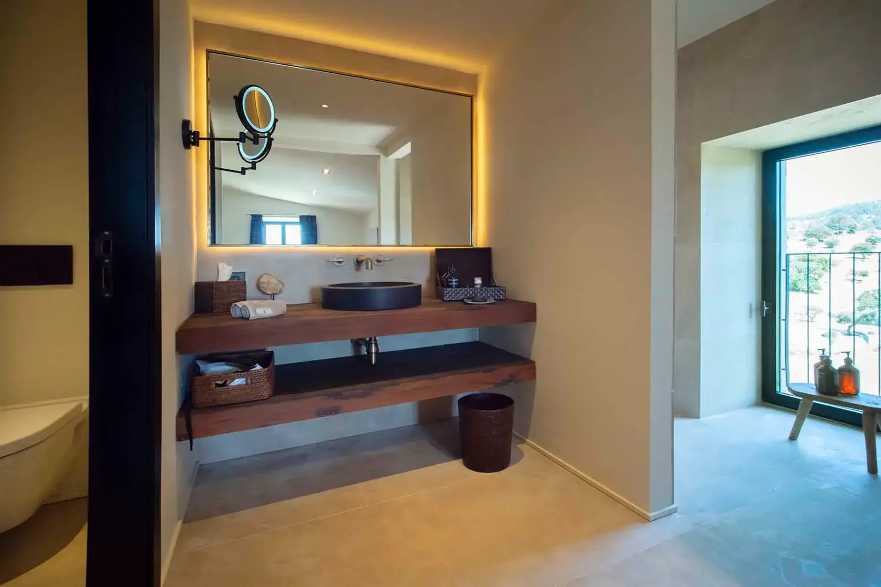 Modern bathroom with large mirror, backlit lighting, wooden shelves, round black sink, and a scenic window view. Neutral color scheme.