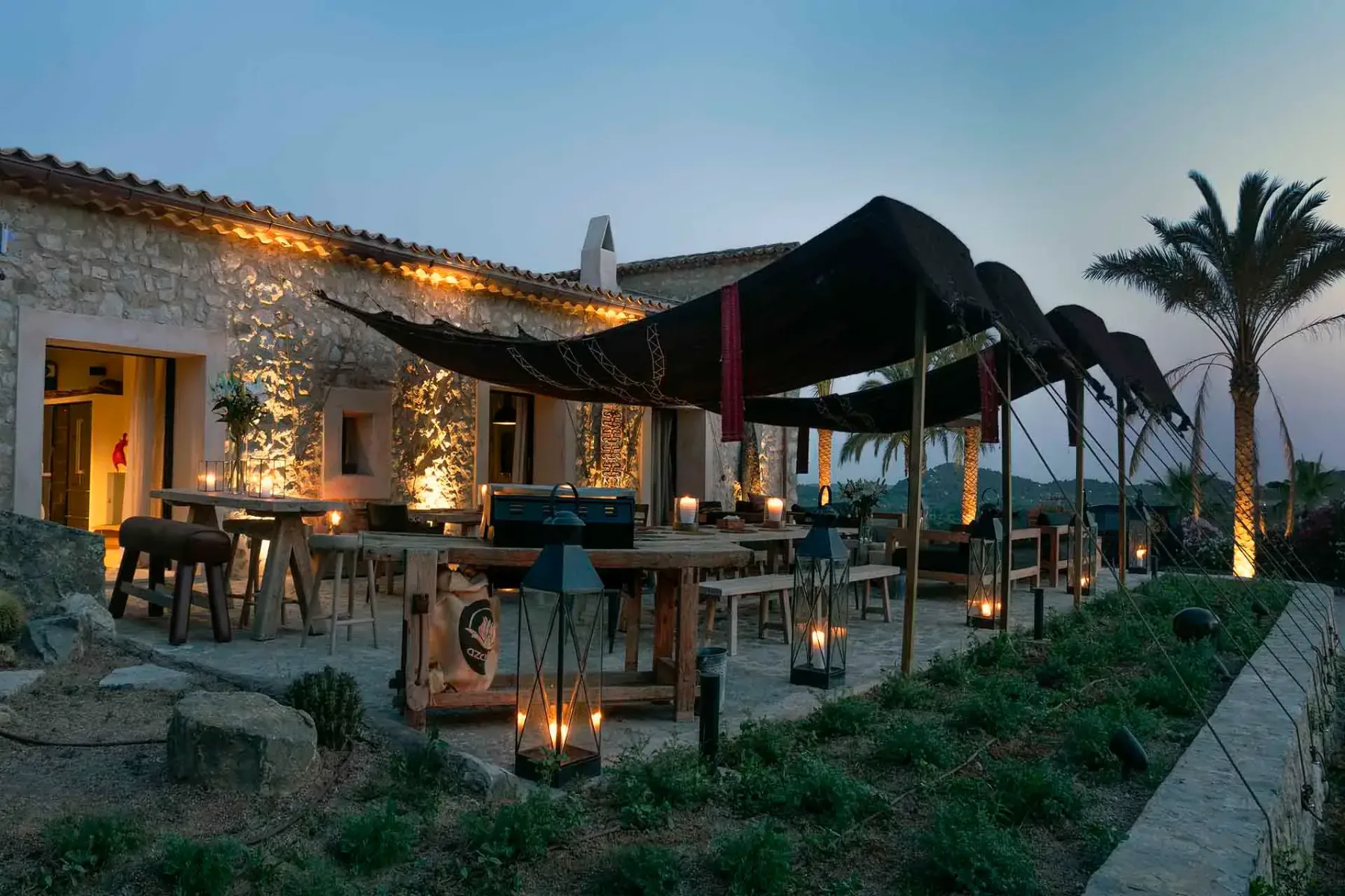 Rustic outdoor restaurant at dusk with stone walls, wooden tables, and lanterns. Draped fabric canopy and palm trees enhance the ambiance.