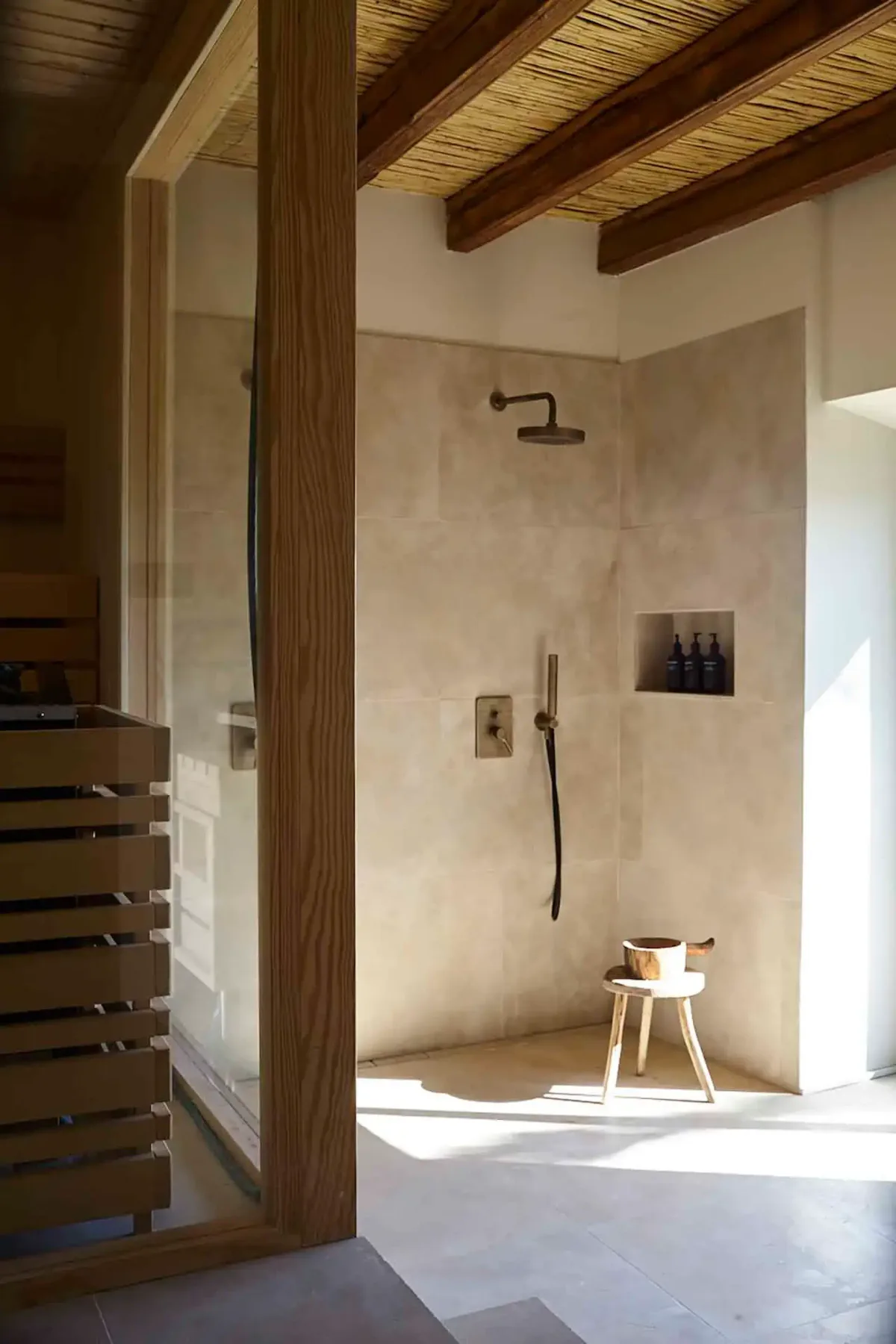 Minimalist bathroom with stone tile, wooden beams, a walk-in shower, and a small wooden stool. Natural light streams through a nearby window.