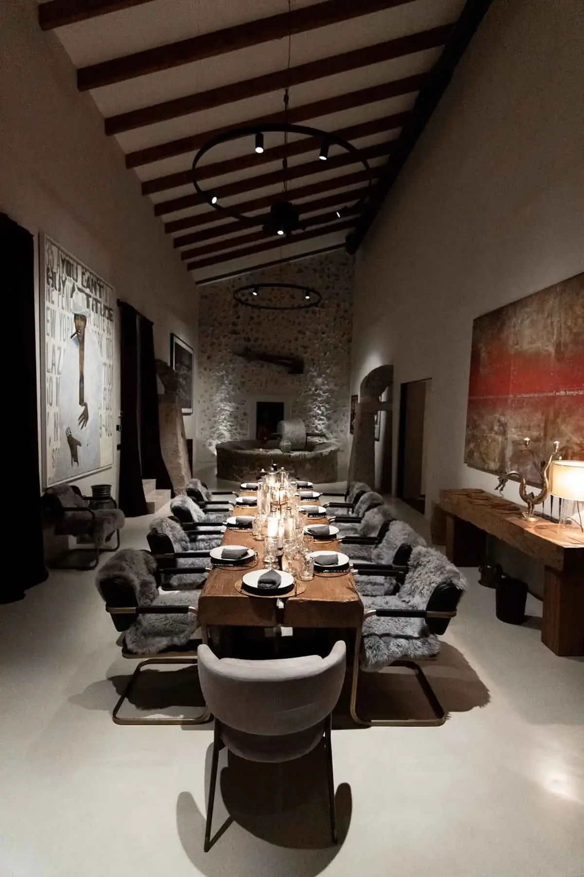 A dimly lit, modern dining room with a long wooden table, set with plates and candles, surrounded by cushioned chairs and large wall art.