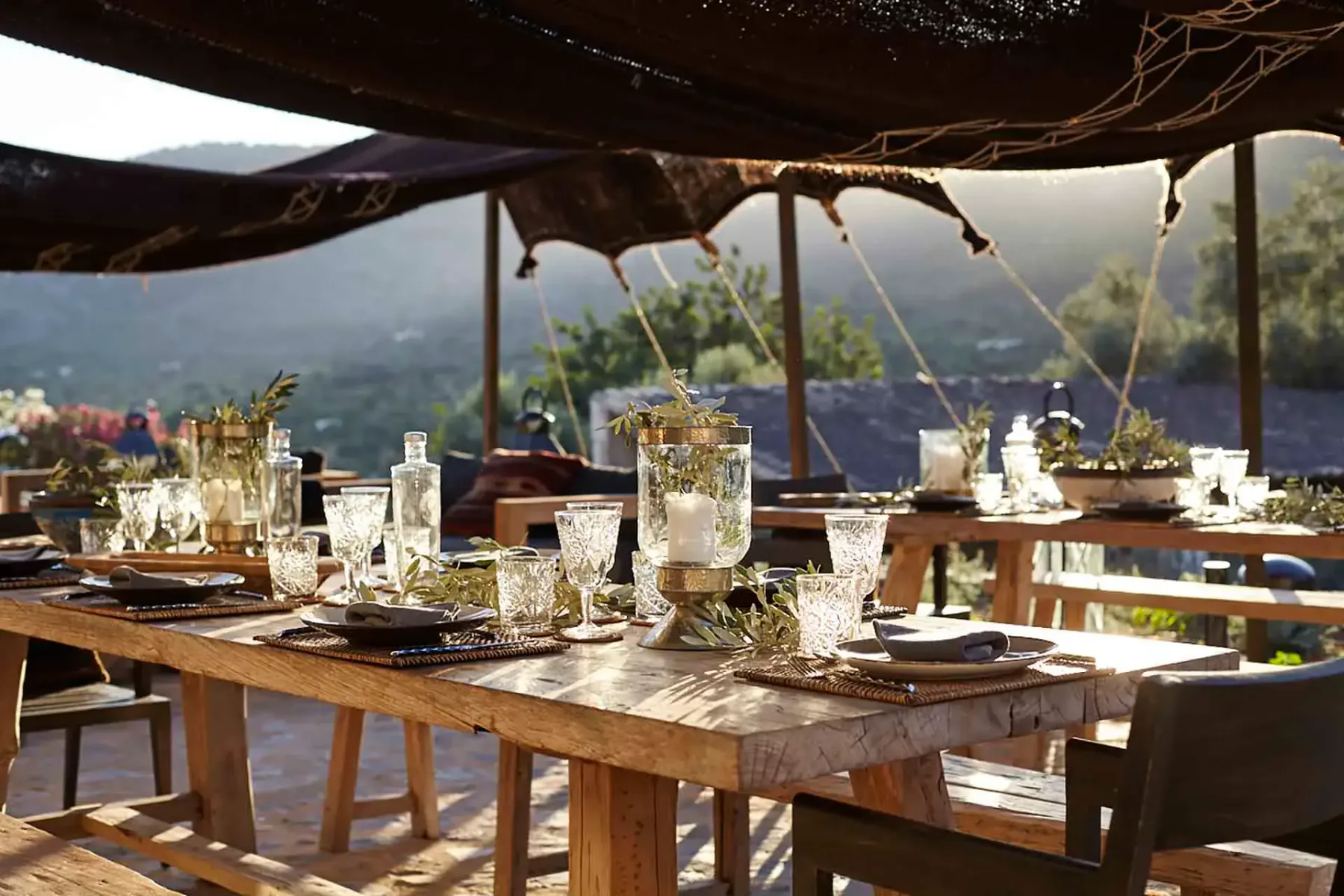 A rustic outdoor dining setup with wooden tables, crystal glasses, candles, and foliage under a canopy, overlooking hills in daylight.