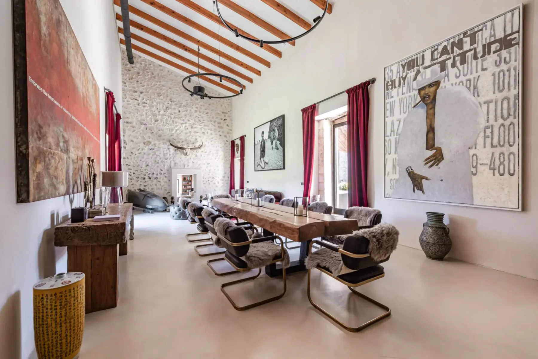 Modern dining room with a long wooden table, fur-covered chairs, large abstract art, stone accent wall, and red curtains. Natural light streams in.