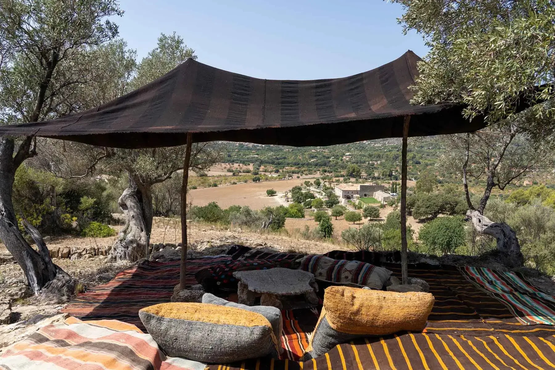 A shaded area with rustic rugs and cushions under a canopy, overlooking a rural landscape with fields and distant buildings.