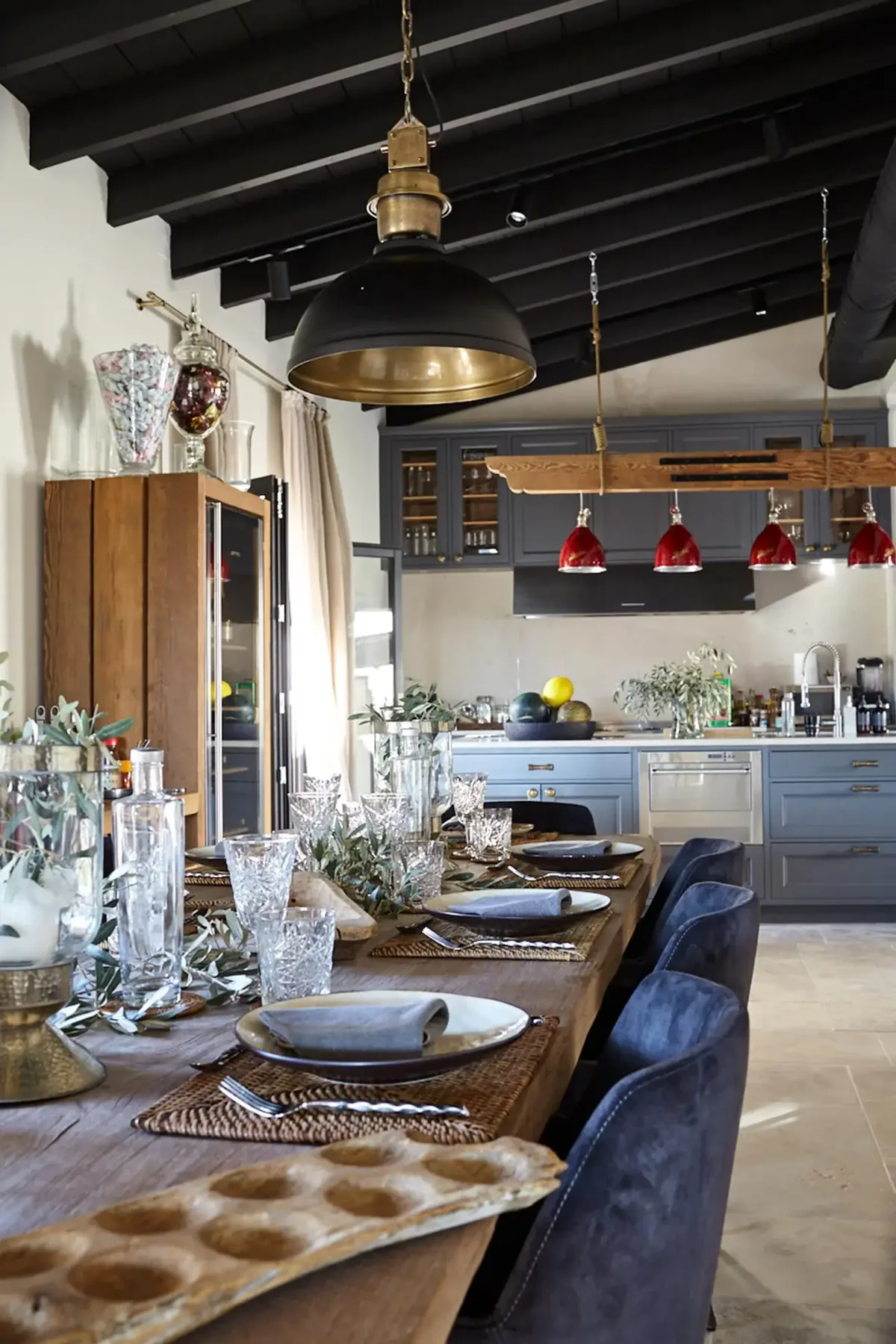 Rustic kitchen with a wooden dining table, elegant glassware, blue cabinets, red pendant lights, and exposed beams on the ceiling.