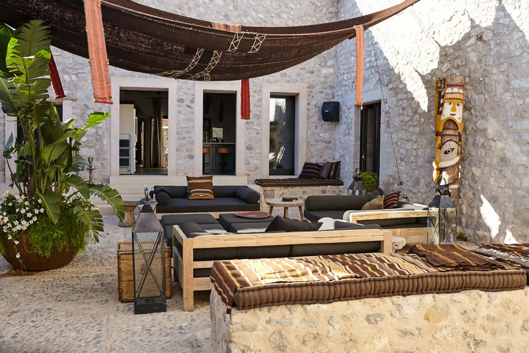 Outdoor patio with stone walls, wooden seating, cushions, and a canopy. Decor includes a large plant and vibrant tapestry.