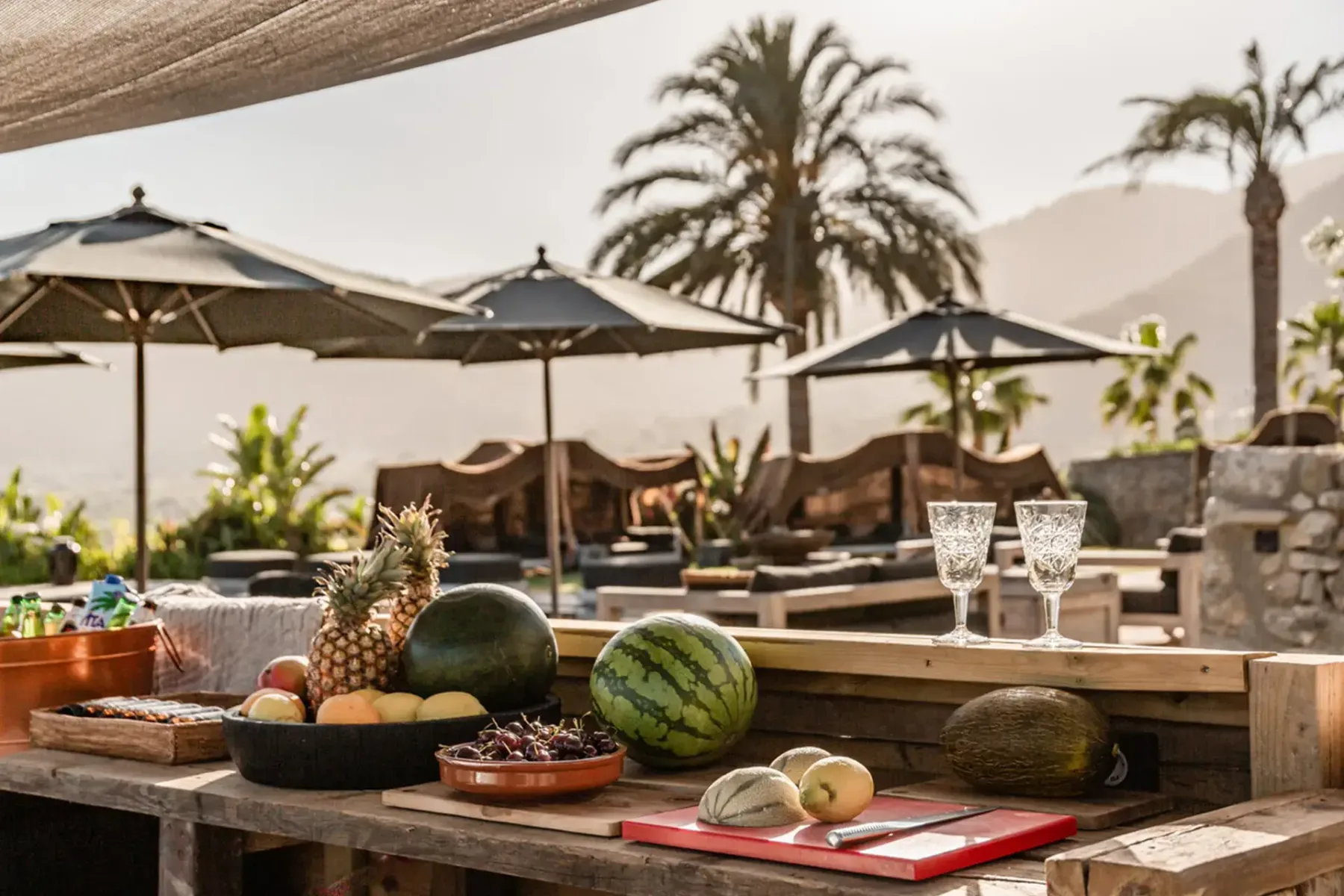 Outdoor buffet with fruit, glasses, and snacks on a wooden table. Umbrellas and palm trees in the background under a sunny sky.