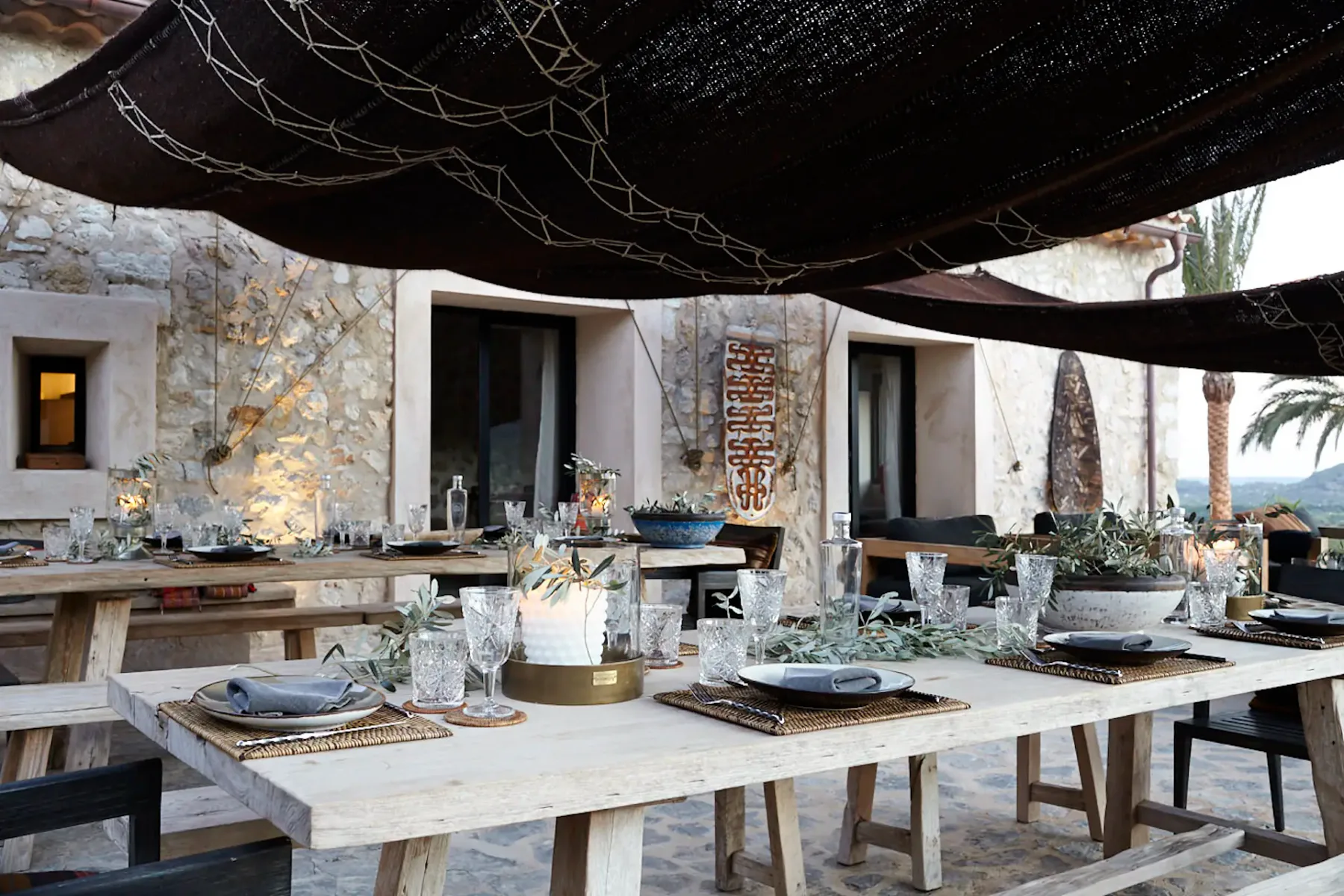 Rustic outdoor dining area with wooden tables set with plates, glasses, and olive branches, under woven canopies on a stone terrace.