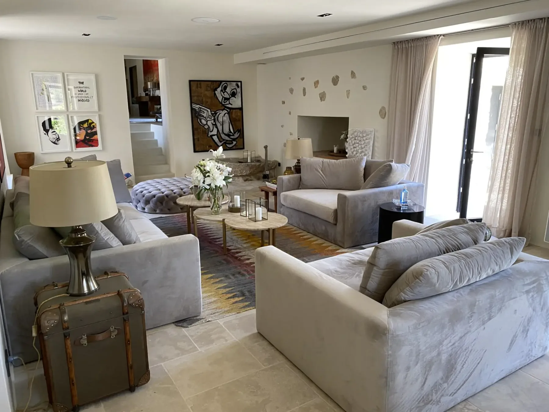 Modern living room with light gray sofas, patterned rug, art on walls, glass coffee tables, and a suitcase-style side table near an open door.