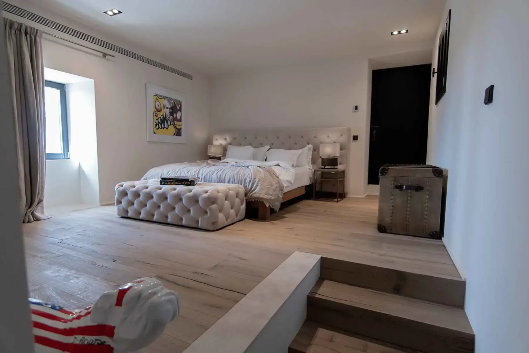 Modern bedroom with a raised platform, tufted bed, upholstered bench, abstract art, wooden floor, and soft lighting.