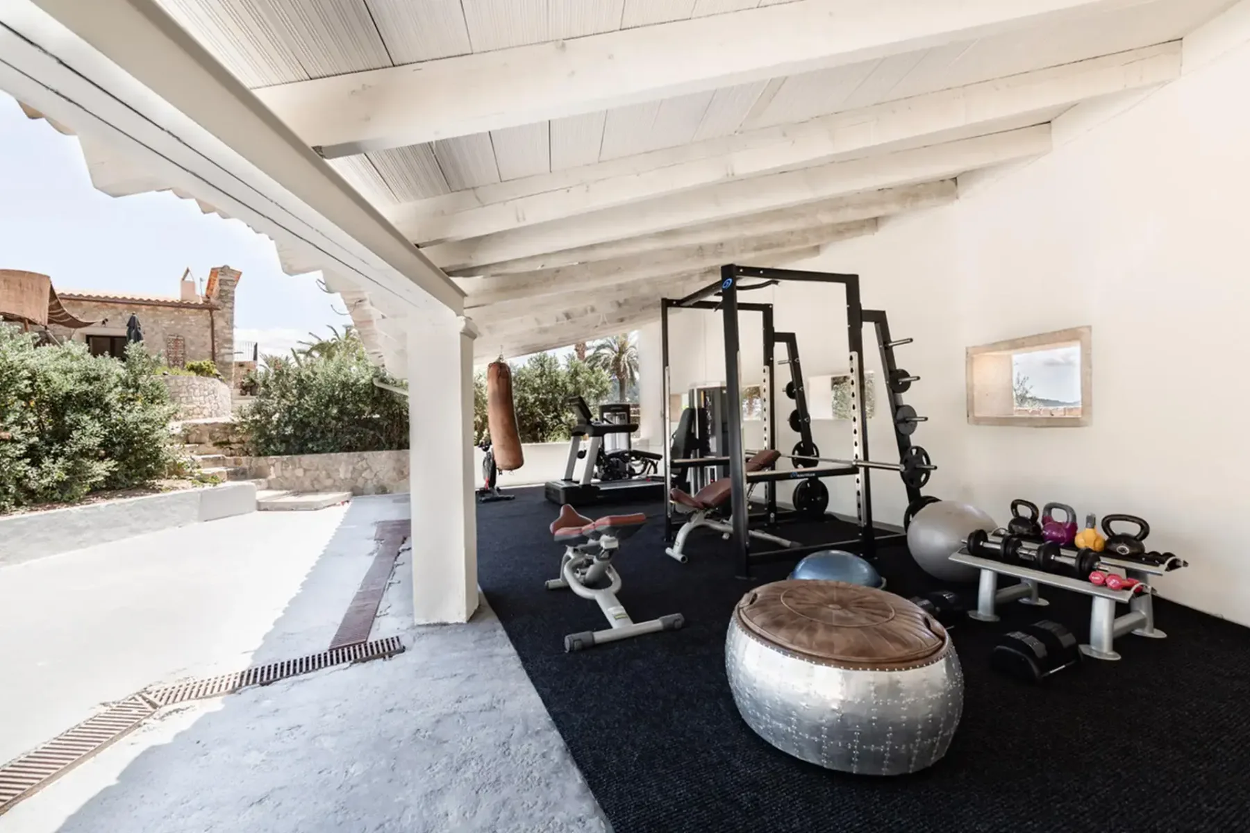 Outdoor home gym with weights, exercise equipment, boxing bag, and yoga ball under a covered patio, overlooking a garden area.