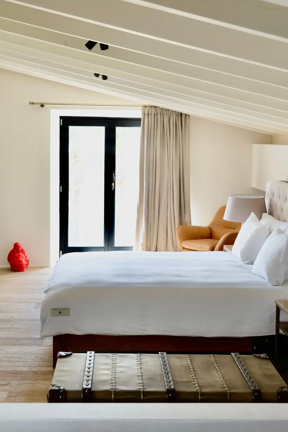 Cozy bedroom with slanted ceiling, double bed, white bedding, large window with beige curtains, an orange chair, and a red decorative figure.
