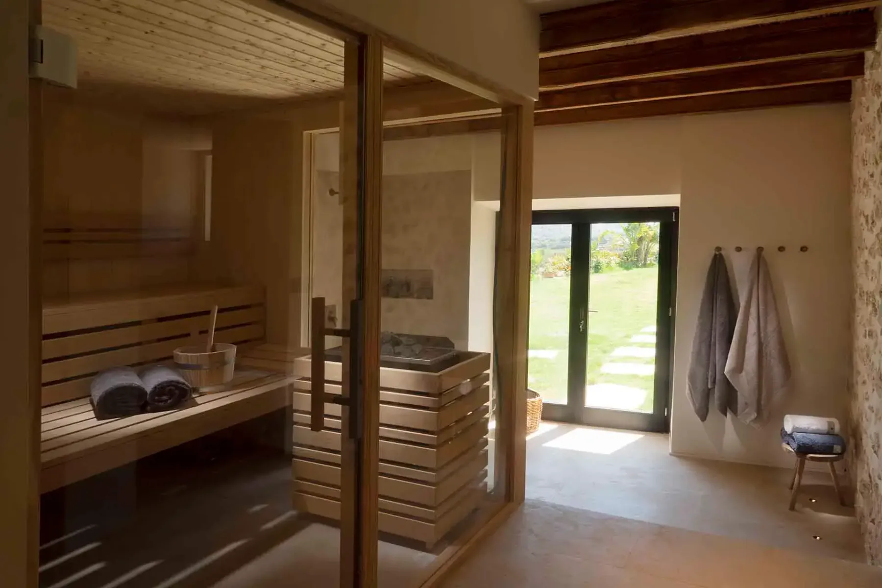 A cozy indoor sauna with wood interiors, towels, and a bucket. Sunlit room with stone wall and a door opening to a garden.