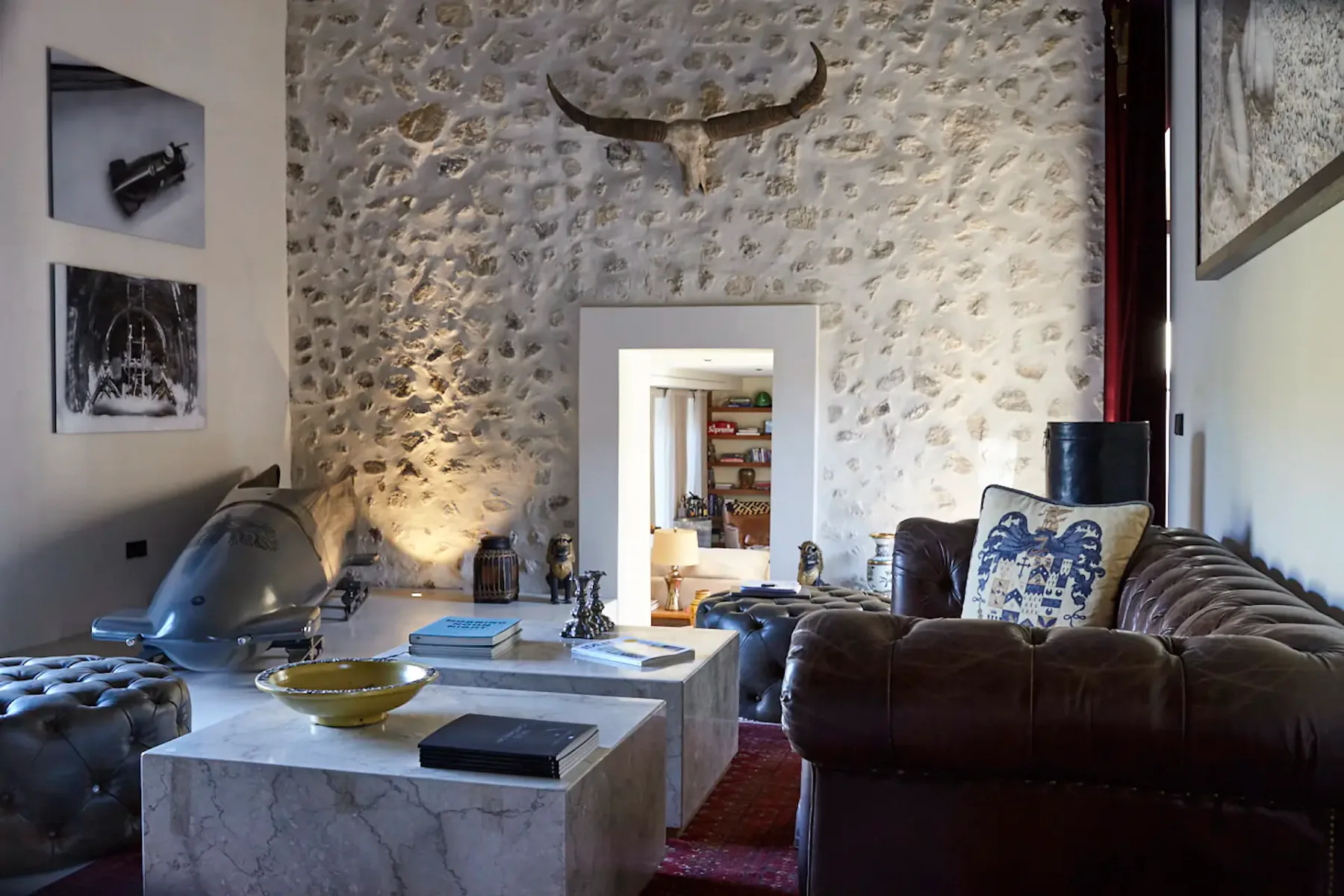 Rustic living room with stone walls, leather sofas, marble tables, and mounted horns. A doorway leads to a bright adjoining room.