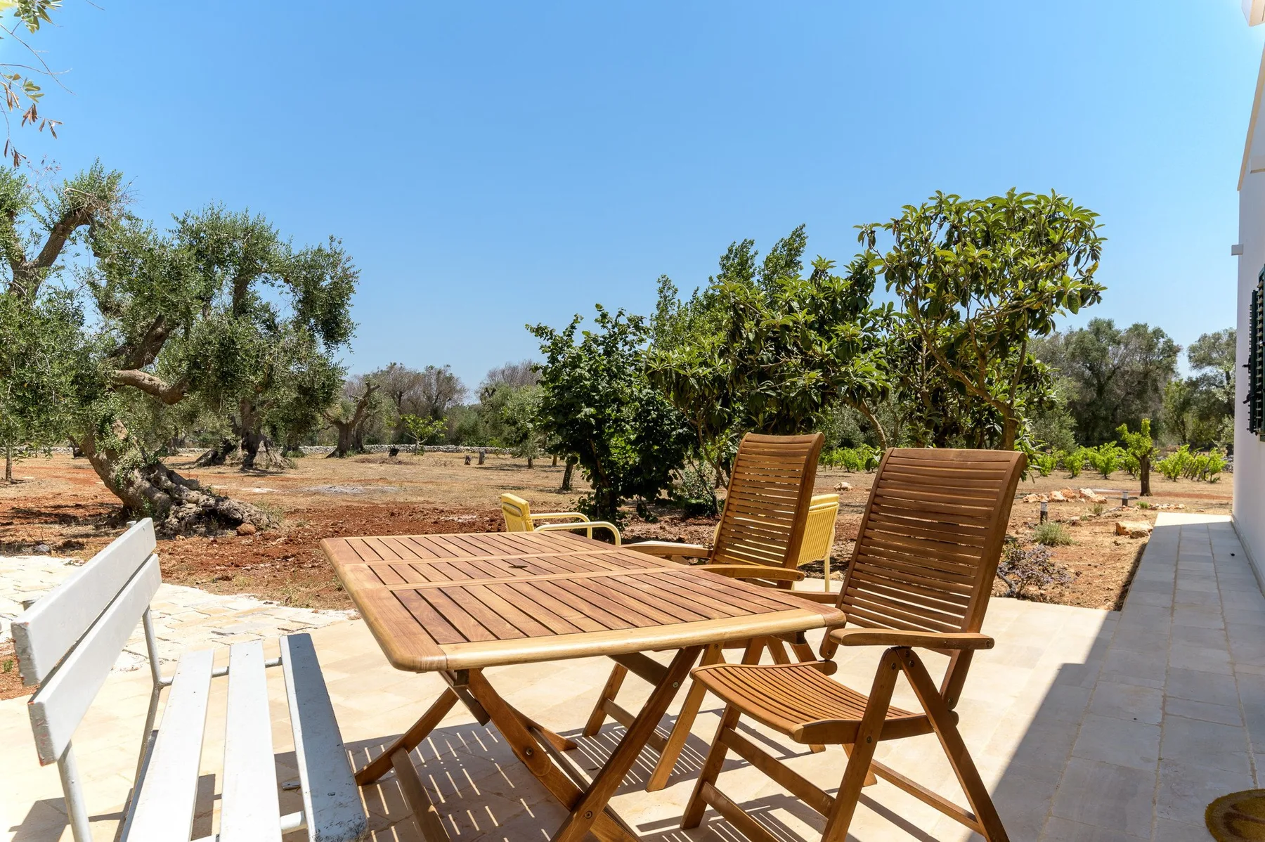 Una mesa de patio de madera y asientos están junto a un jardín bajo un cielo despejado.
