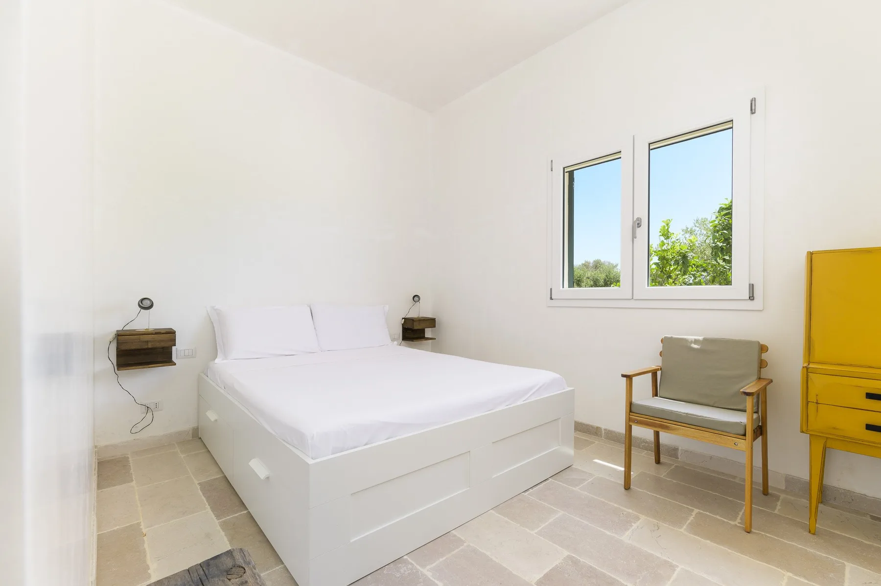 Un dormitorio luminoso con una cama blanca, una ventana y asientos sencillos.