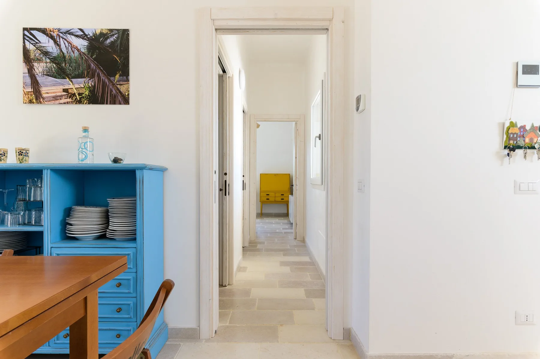 Un pasillo luminoso pasa junto a un mueble azul y conduce hacia una silla amarilla.