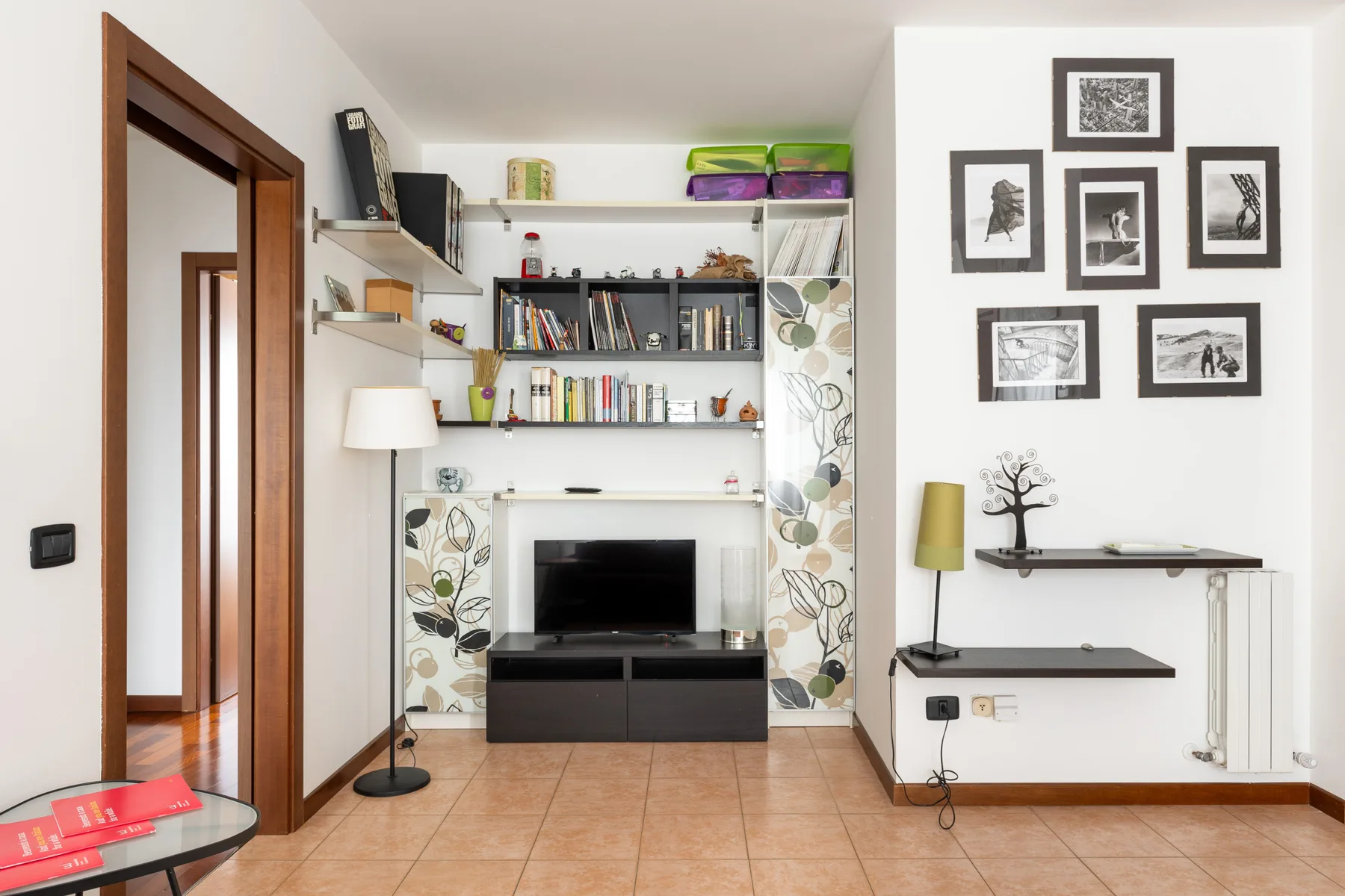 Una sala luminosa tiene un televisor bajo estantes de pared y cuadros enmarcados.