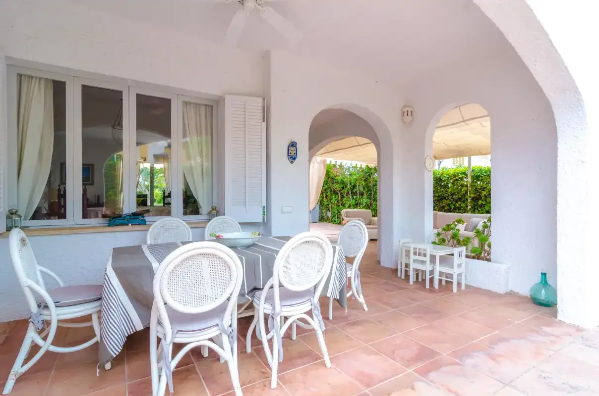 A covered patio with a dining table and white seating beside arched openings.