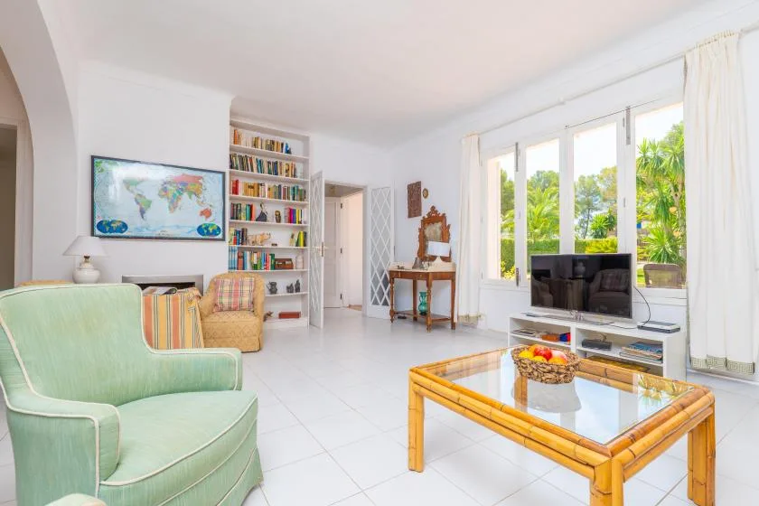A bright living room with seating, a television, and large windows overlooking greenery.