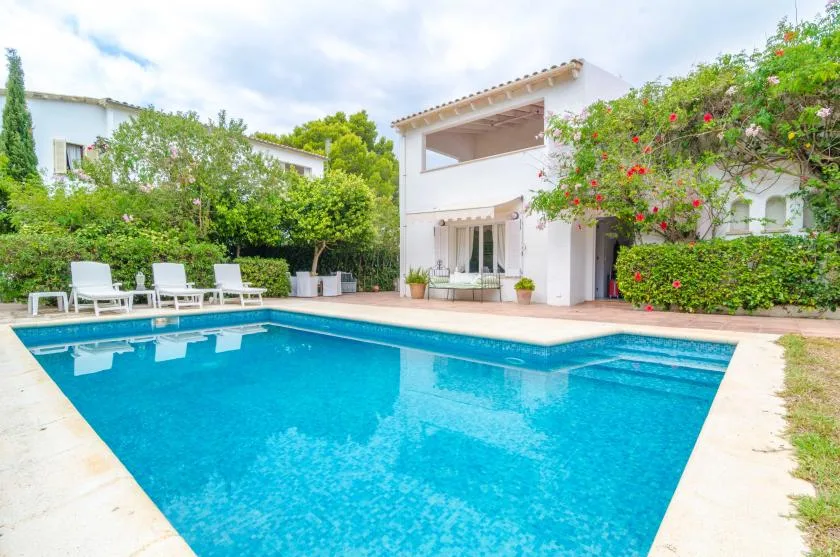An outdoor swimming pool beside a white house and leafy garden.