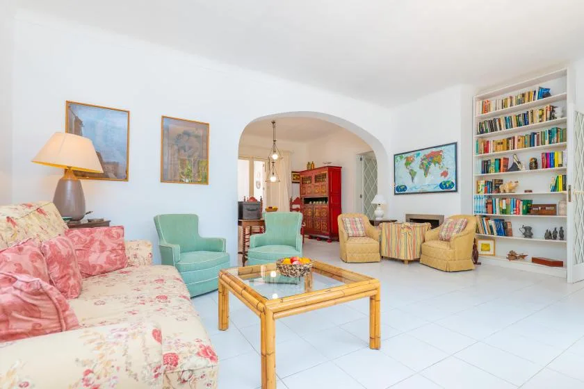 A bright living room with patterned seating, a glass coffee table, and a wall of bookshelves.