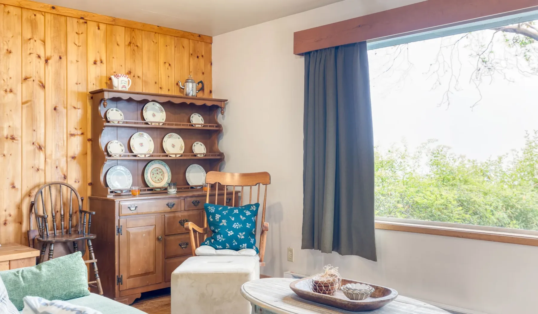 Una sala luminosa con un mueble de madera que exhibe vajilla junto a una ventana grande.
