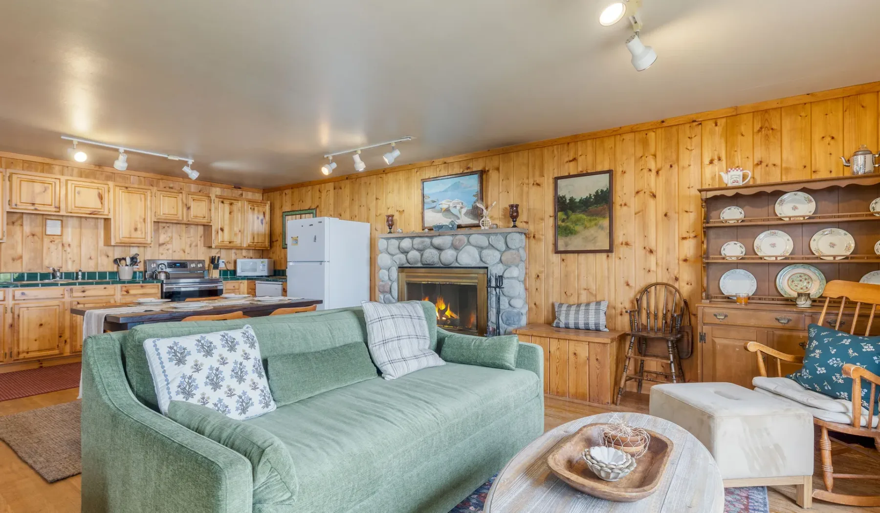 Una sala con paredes de madera se abre a una cocina, con asientos verdes frente a una chimenea de piedra.