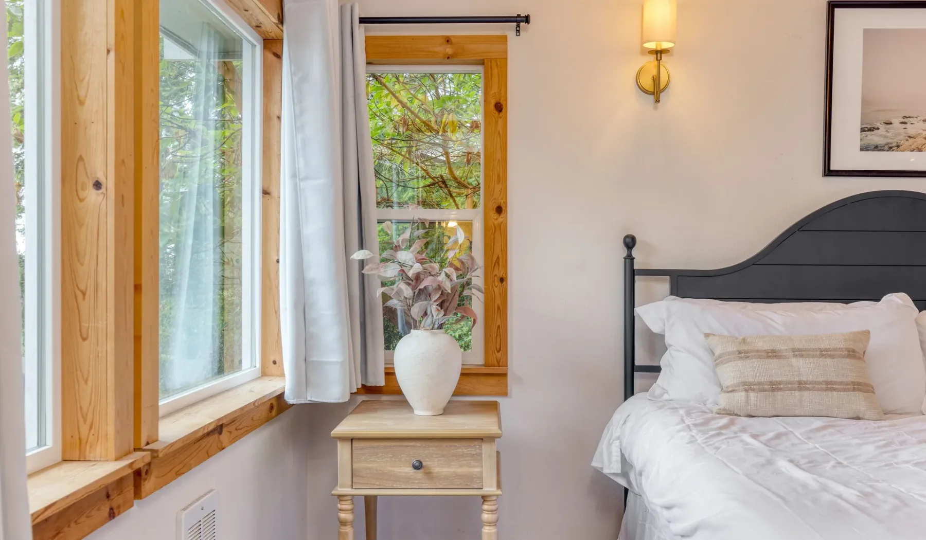 Un dormitorio con una cama junto a ventanas con marcos de madera y una mesita de noche.