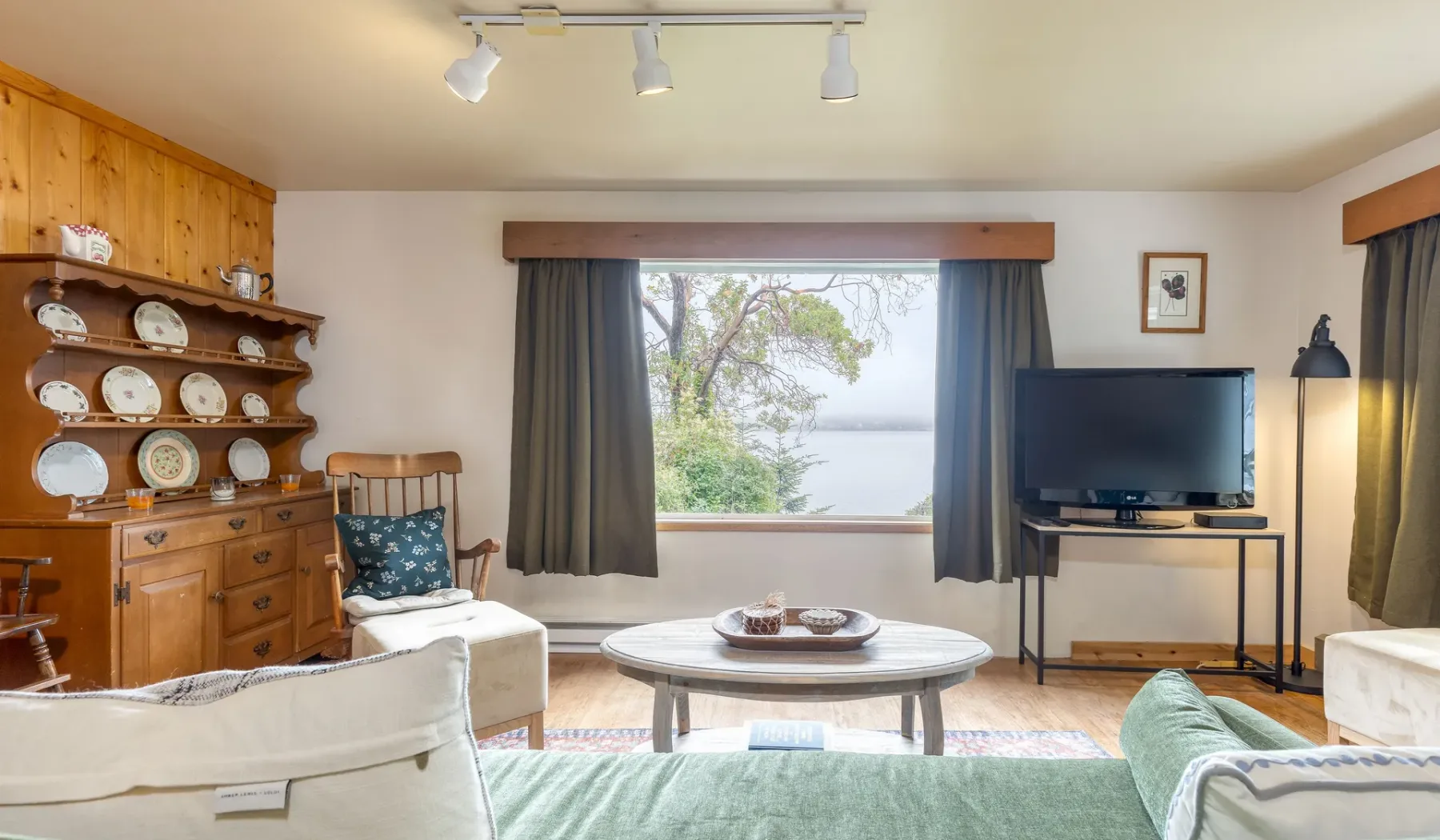 Una sala de estar con asientos, un televisor y una ventana amplia con vista a árboles y agua.