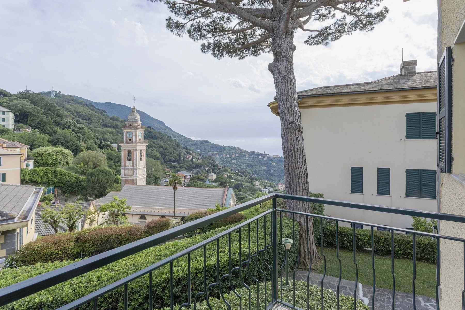 A balcony overlooks a hillside town, with a bell tower and tree in view.