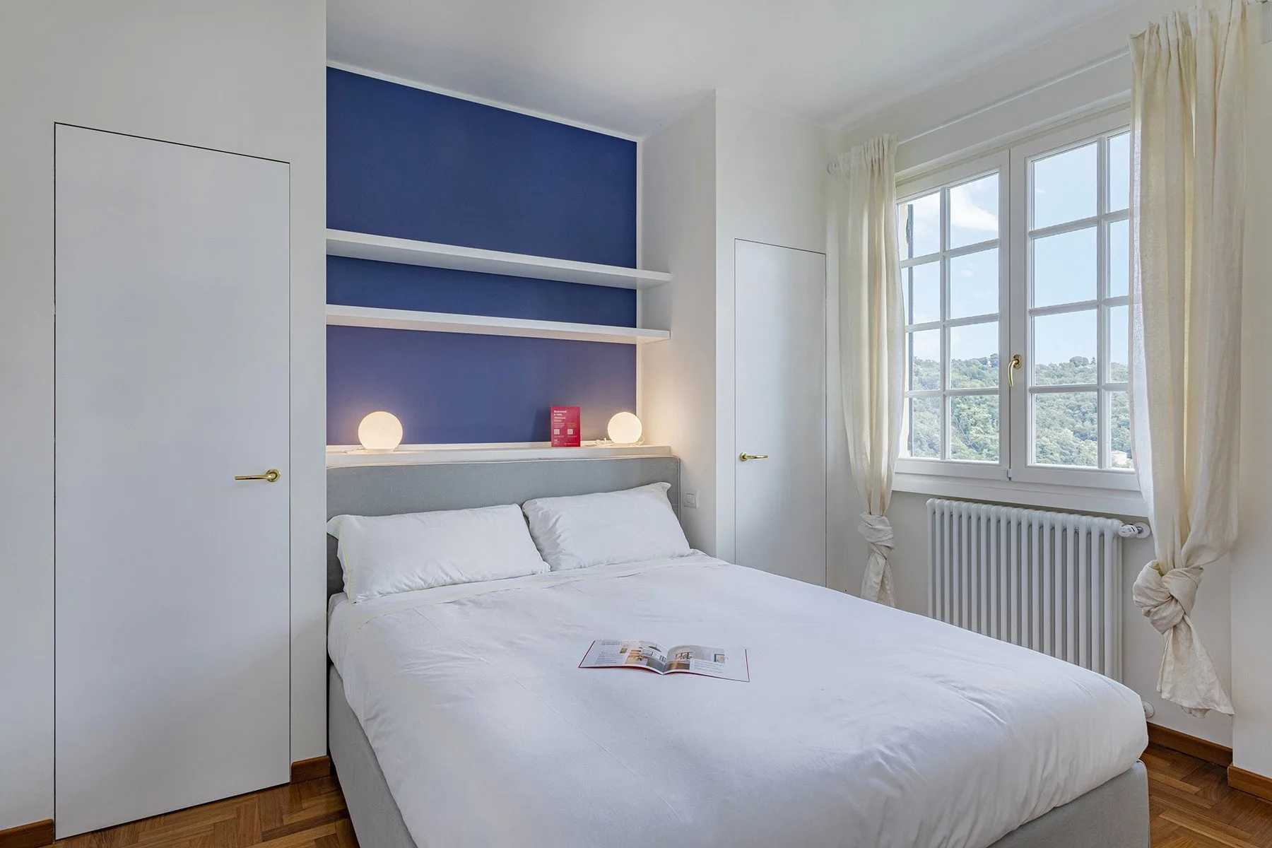 A bedroom with a bed beneath blue wall shelving and a large window.