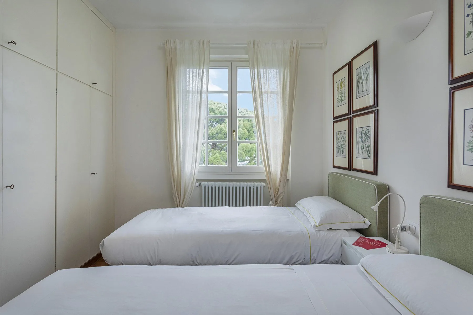 A bright bedroom with two beds, a window, and framed artwork.