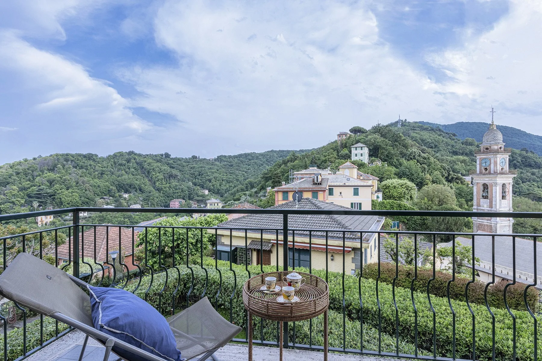 A terrace overlooks green hills and a church tower, with outdoor seating by the railing.