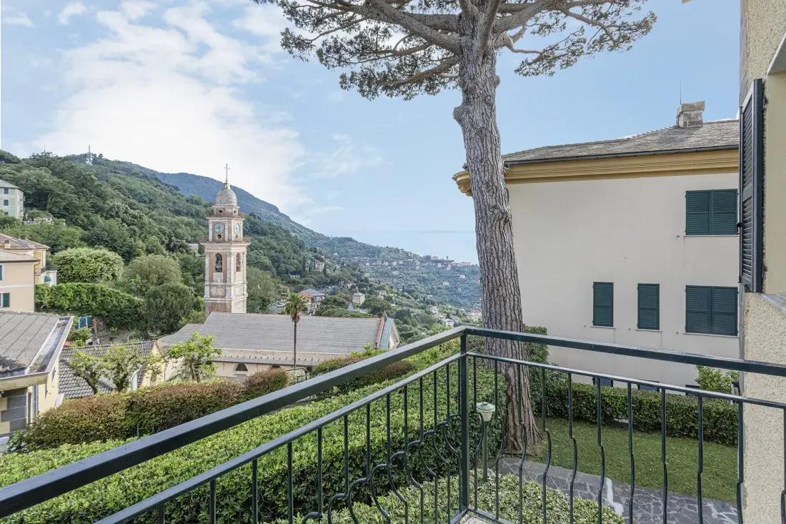 A balcony overlooks a hillside town, with a church tower and tree in view.
