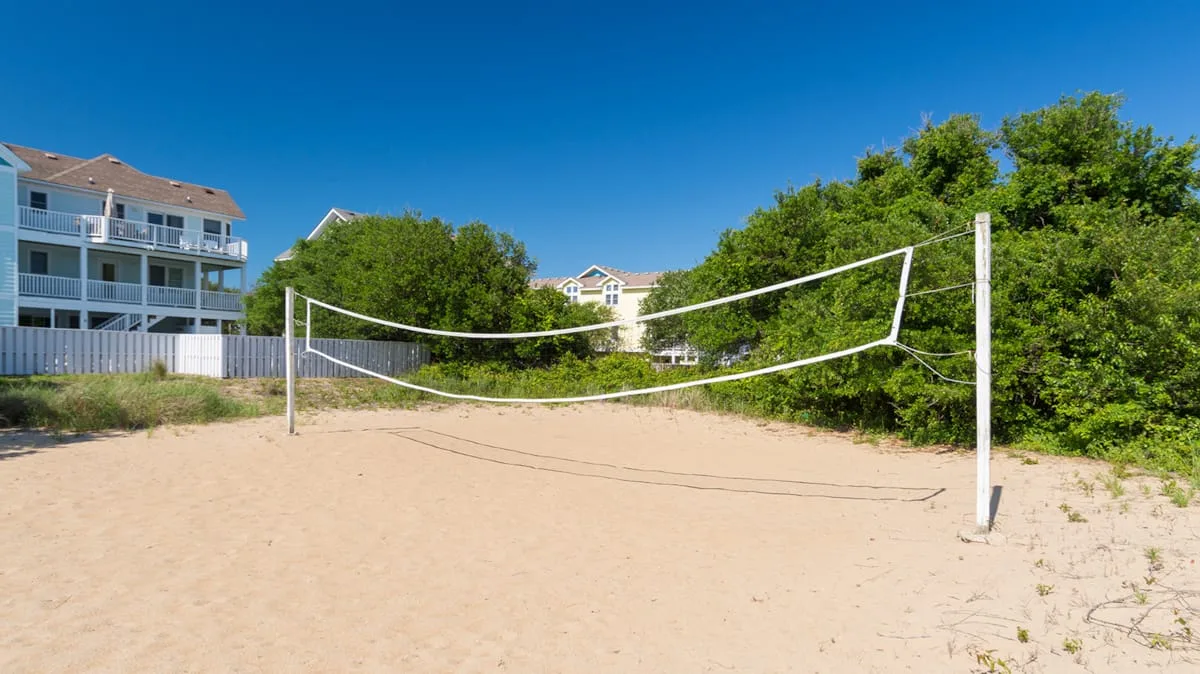 A sandy outdoor volleyball court is set beside trees and nearby buildings.