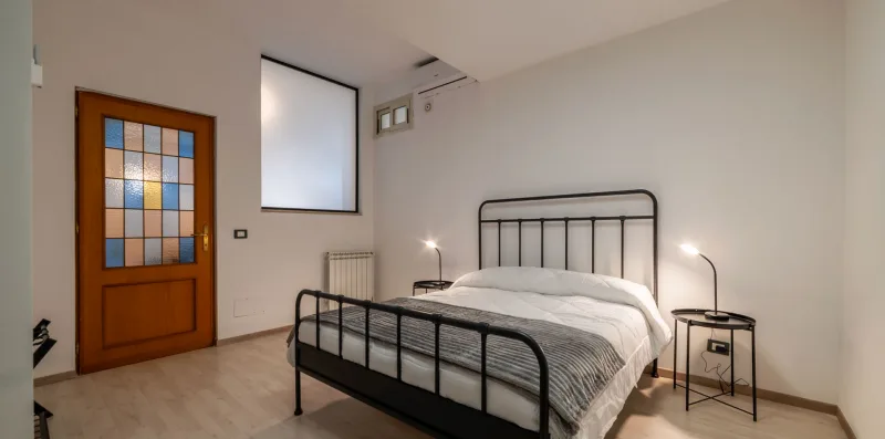 Un dormitorio con una cama de metal, ropa de cama estampada y una ventana con marco de madera.