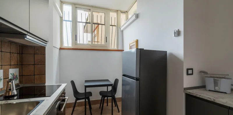 Una cocina con una mesa de comedor junto a un refrigerador alto y ventanas luminosas.