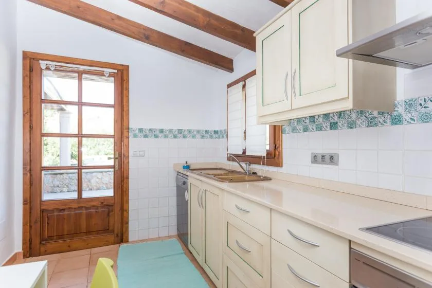 A bright kitchen with white cabinets, a long counter, and exposed wooden ceiling beams.