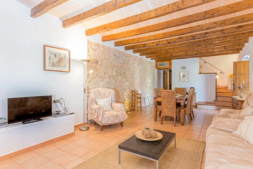 A bright living and dining area with exposed wooden ceiling beams and a television.