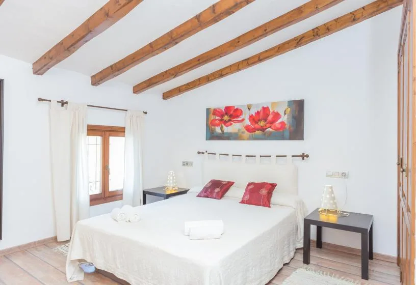A bright bedroom with a white bed, red pillows, and exposed wooden ceiling beams.